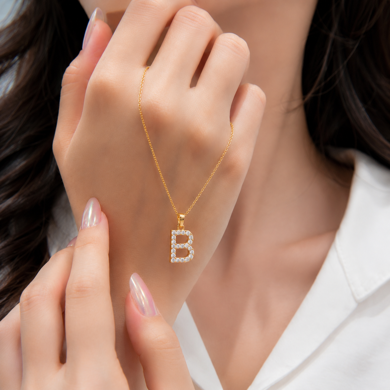A close-up lifestyle shot of a model wearing a delicate gold initial necklace with a letter 'B' pendant encrusted with small clear stones. The model is wearing a white collared shirt, and her hands are styled elegantly near her face.